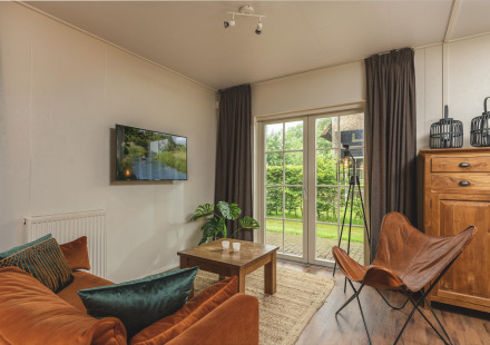 Knusse glamping woonkamer met oranje bank, vlinderstoel, houten meubels en uitzicht op de groene tuin.