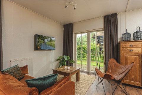Sala de glamping acogedora con sofá naranja, silla mariposa y vistas al jardín verde a través de puertas de cristal.