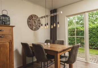 Moderne eetruimte bij glamping, met houten tafel, vier stoelen, grote klok en tuinuitzicht door ramen.