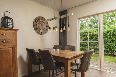 Moderne eetplek in glamping-accommodatie met houten tafel, vier stoelen, grote klok en zicht op tuin.