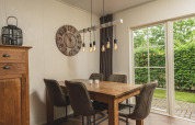 Modern dining area at glamping site with wooden table, four chairs, large wall clock and garden view windows.