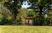 Kleine Holzhütte mit Terrasse zwischen Bäumen im Holiday Park Mölke, Niederlande, umgeben von Natur.