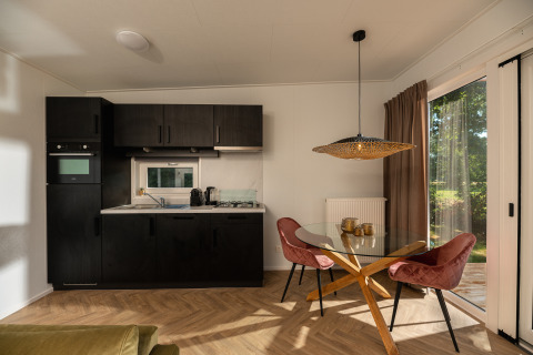 Moderne tiny house met zwarte keuken, glazen eetkamertafel, roze stoelen en zicht op de natuur.