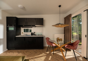 Moderne tiny house met zwarte keuken, glazen eetkamertafel, roze stoelen en zicht op de natuur.