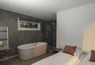 Moderne glampingkamer met losstaand bad, douche, comfortabel bed en groot raam met uitzicht op natuur.