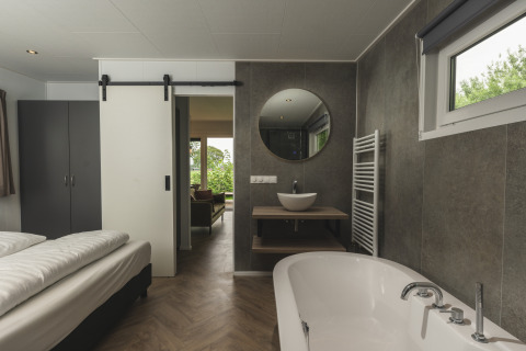 Moderne glamping accommodatie met slaapkamer, open badkamer, vrijstaand bad en uitzicht op groen.
