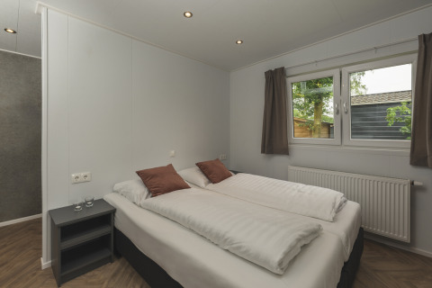 Moderne glamping slaapkamer met dubbel bed, bruine kussens, nachtkastje en raam met zicht op natuur.