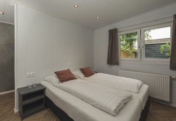 Moderne glamping slaapkamer met dubbel bed, bruine kussens, nachtkastje en raam met zicht op natuur.