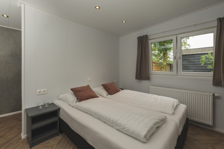 Dormitorio de glamping moderno con cama doble, cojines marrones, mesa lateral y ventana hacia la naturaleza.