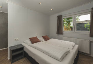 Moderne glamping slaapkamer met tweepersoonsbed, bruine kussens, nachtkastje en raam met uitzicht op groen.