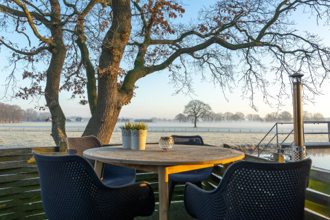 Runder Holztisch mit vier Stühlen auf Terrasse, Blick auf Wiesen und Bäume im Regge Cottage, Niederlande.