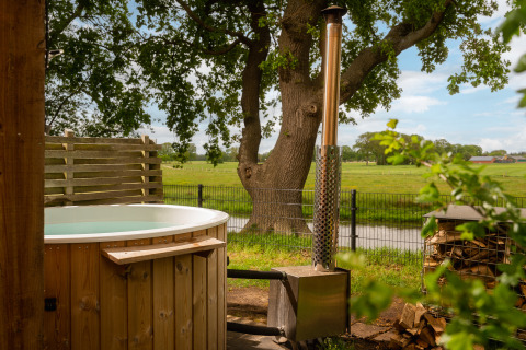 Udendørs træ-spa med brændeovn ved Regge Cottage på Holiday Park Mölke, omgivet af grønne marker.