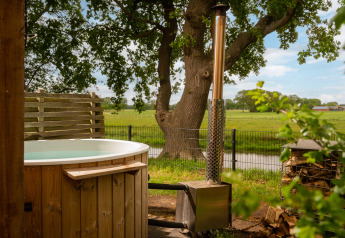 Udendørs træ-spa med brændeovn ved Regge Cottage på Holiday Park Mölke, omgivet af grønne marker.