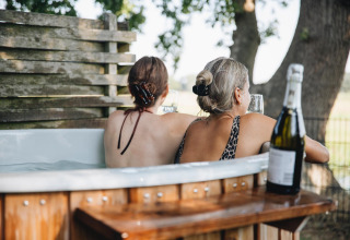 Two women relax in a hot tub with wine at Regge Cottage tiny house in Holiday Park Mölke, Netherlands.