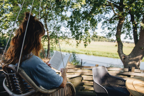 Kvinde læser bog i en hængestol på terrassen med udsigt over flod og natur ved Holiday Park Mölke.