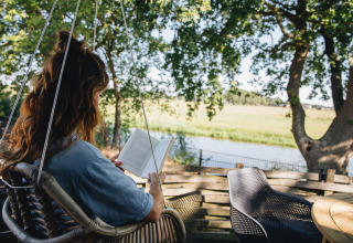 Kvinde læser bog i en hængestol på terrassen med udsigt over flod og natur ved Holiday Park Mölke.