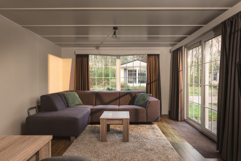 Sala de estar con sofá grande y ventanales en Bungalette Basic + hidromasaje en Holiday Park Mölke, Países Bajos.