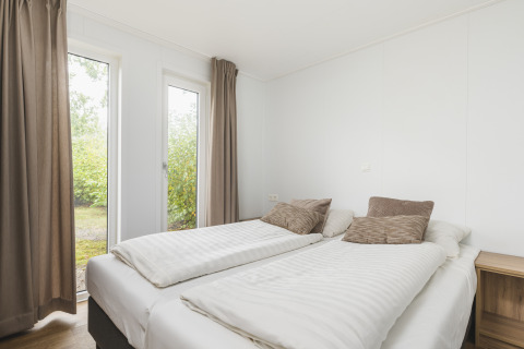 Dormitorio de glamping luminoso con dos camas individuales, cortinas beige y vista al jardín exterior.