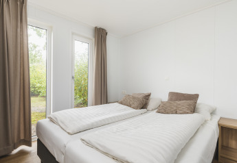 Bright glamping accommodation bedroom with twin beds, beige curtains, and garden view through windows.