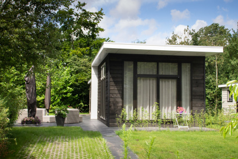 Et moderne, sort træhytte med store vinduer omgivet af grønne træer og hyggelig terrasse i solen.