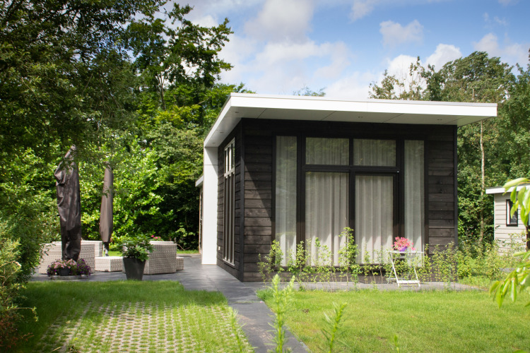 Cabaña moderna de madera oscura con ventanas grandes, jardín exuberante y terraza sombreada y tranquila.