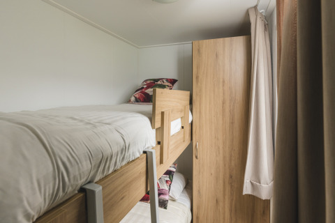 Petite chambre glamping avec lits superposés en bois, linge blanc, armoire, et rideaux beiges.