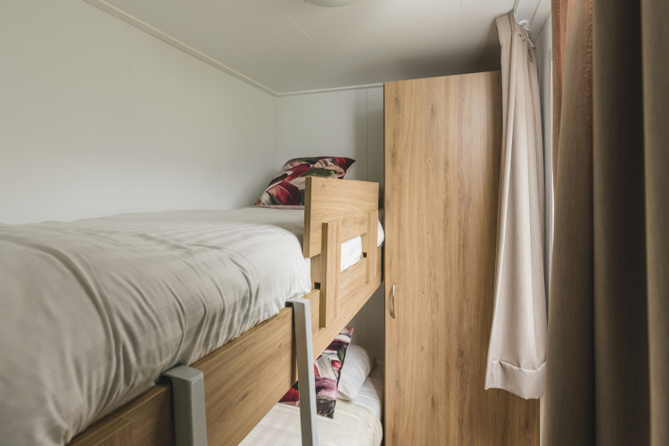 Habitación glamping pequeña con literas de madera, ropa de cama blanca, armario y cortinas claras.