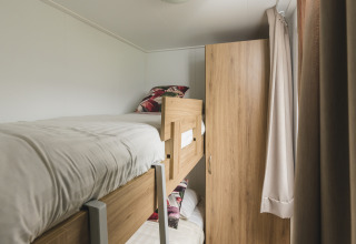 Compacte glampingkamer met houten stapelbedden, witte lakens, kast en lichte gordijnen.