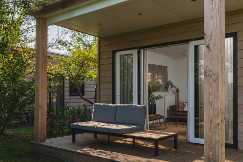Terraza exterior de una moderna cabaña con sofá y mesa, rodeada de vegetación y puerta abierta al salón.
