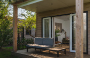 Outdoor deck of a modern lodge with a sofa and table, surrounded by greenery, doors open to living room.