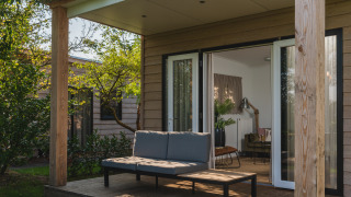 Outdoor deck of a modern lodge with a sofa and table, surrounded by greenery, doors open to living room.