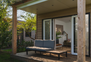 Terrasse einer modernen Lodge mit Sofa und Tisch, umgeben von Bäumen, Blick ins gemütliche Wohnzimmer.