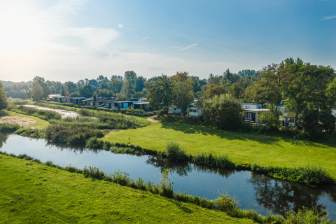 Fluss-Lodges im Holiday Park Mölke, Niederlande, mit grünen Wiesen, Bäumen und ruhigem Wasser im Sonnenschein.
