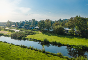 Fluss-Lodges im Holiday Park Mölke, Niederlande, mit grünen Wiesen, Bäumen und ruhigem Wasser im Sonnenschein.
