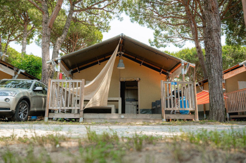 Safari tent Villatent Outback with porch, car parked beside, surrounded by trees on a sunny day.