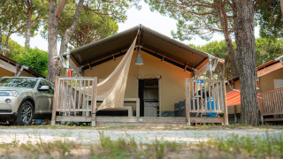 Safari-Zelt Villatent Outback mit Veranda und Auto daneben, umgeben von Bäumen an einem sonnigen Tag.