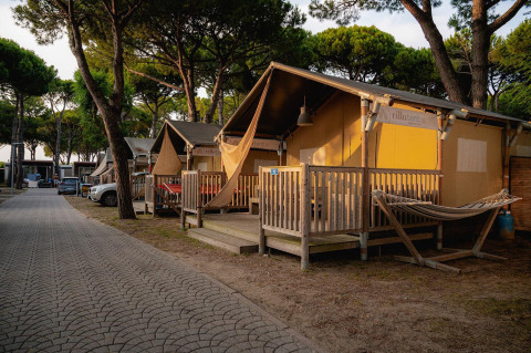 Foto delle tende safari Villatent Outback tra pini, con amaca e veranda in legno illuminate dal tramonto.