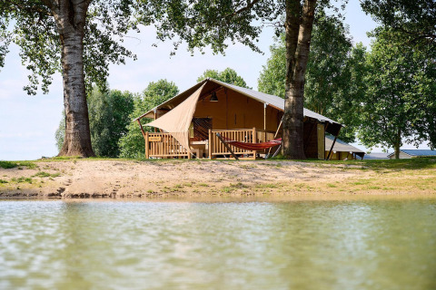 Tienda safari Villatent Nomad en Camping Betuwe, Países Bajos, junto a un lago y rodeada de árboles.