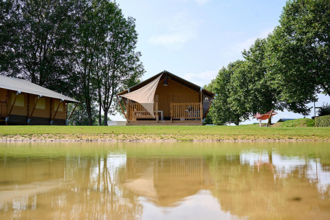Tienda safari Villatent Nomad en Camping Betuwe, Países Bajos, junto a un lago y rodeada de árboles verdes.