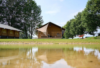 Tienda safari Villatent Nomad en Camping Betuwe, Países Bajos, junto a un lago y rodeada de árboles verdes.