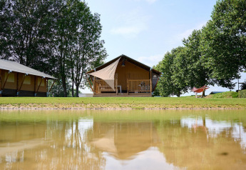 Tienda safari Villatent Nomad en Camping Betuwe, Países Bajos, junto a un lago y rodeada de árboles verdes.