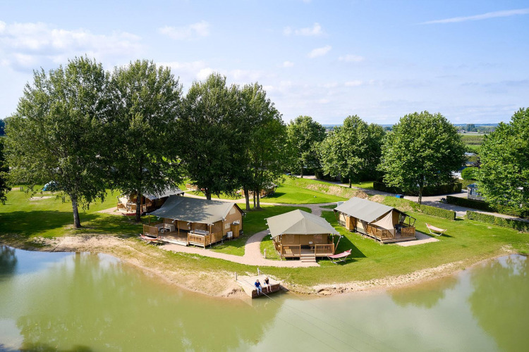 Safari tents by a lakeside surrounded by green trees at Camping Betuwe, Netherlands, ideal for glamping.