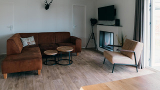 Woonkamer in Veluwe Villa met bruine sofa, moderne zetel, haard, televisie en groot raam naar buiten.