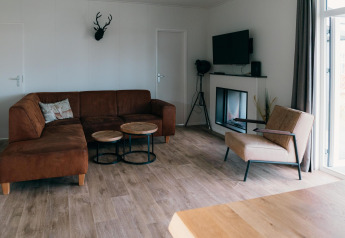 Salon de la Veluwe Villa avec canapé d’angle marron, fauteuil moderne, TV, cheminée et grandes fenêtres.