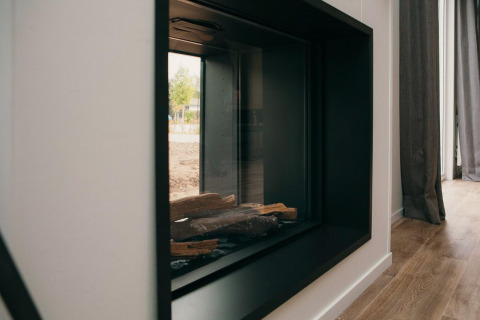 Moderne haard met glas in Veluwe Villa + sauna, houtblokken en reflectie van het buitenzicht zichtbaar.