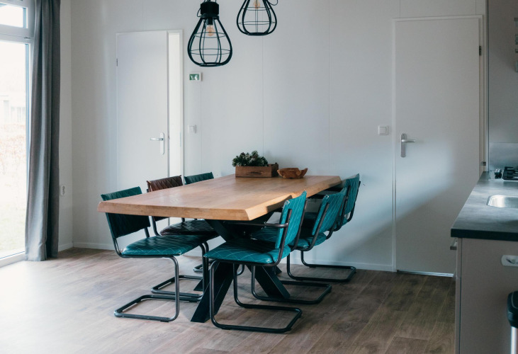 Modern dining area in Veluwe Villa featuring a wooden table, six blue chairs, pendant lights, and sunlight.