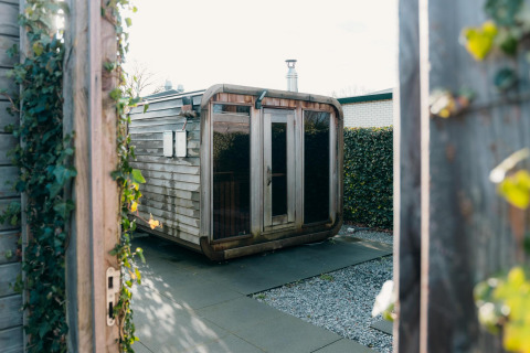Modern wooden sauna at Veluwe Villa, surrounded by hedges and open gates, bathed in natural daylight.