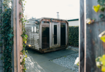 Modern wooden sauna at Veluwe Villa, surrounded by hedges and open gates, bathed in natural daylight.