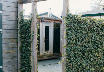 Entrada a la sauna exterior de Veluwe Villa en el parque vacacional De Boshoek, Países Bajos, con setos verdes.