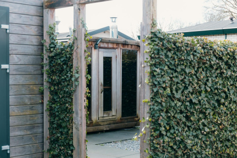 Entrada a la sauna exterior de Veluwe Villa en el parque vacacional De Boshoek, Países Bajos, con setos verdes.